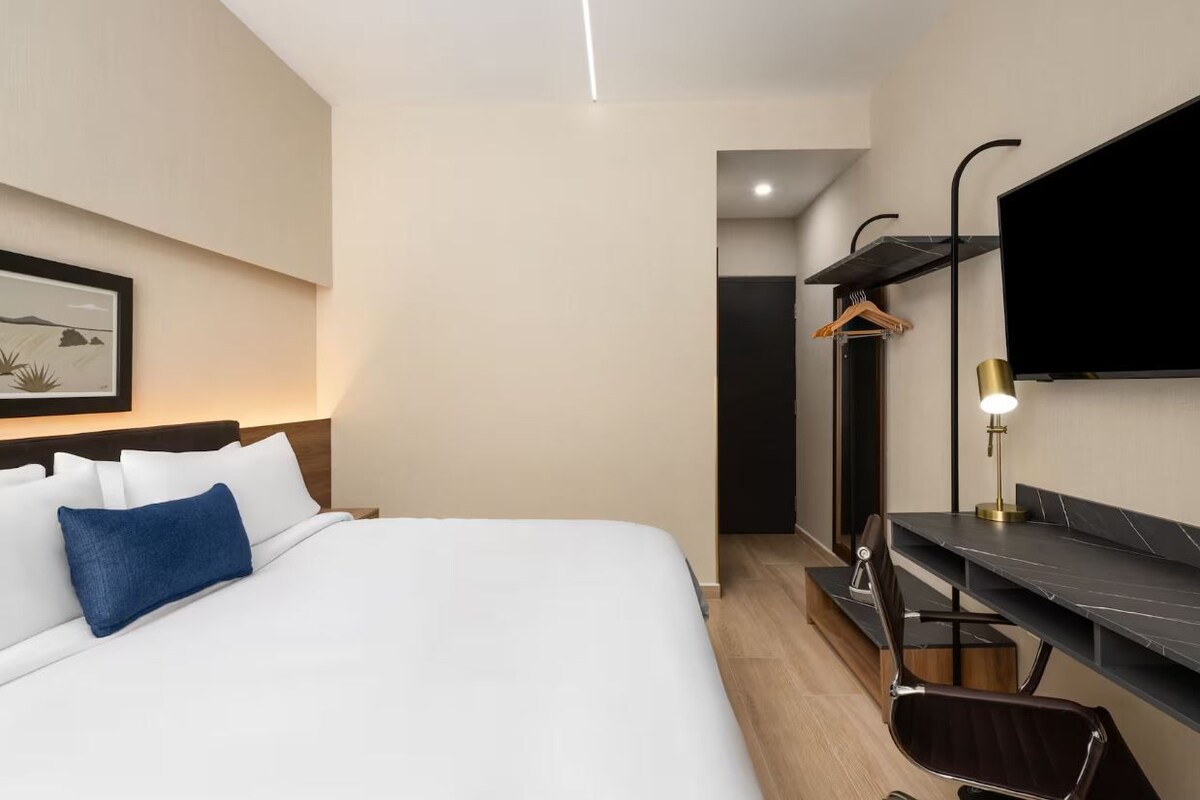 A modern hotel room with a large bed, a desk with a chair, a flat-screen TV mounted on the wall, and a framed picture on the wall.