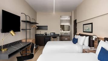 Habitación de hotel con dos camas, un escritorio con computadora, televisor, kitchenette y espejo de baño.