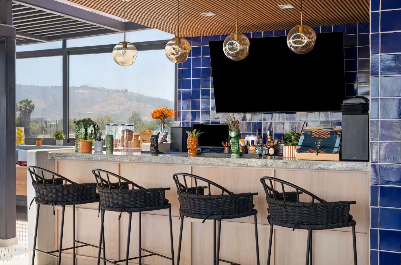 Zona de bar moderna con un televisor grande, taburetes y diferentes botellas y plantas en la barra.