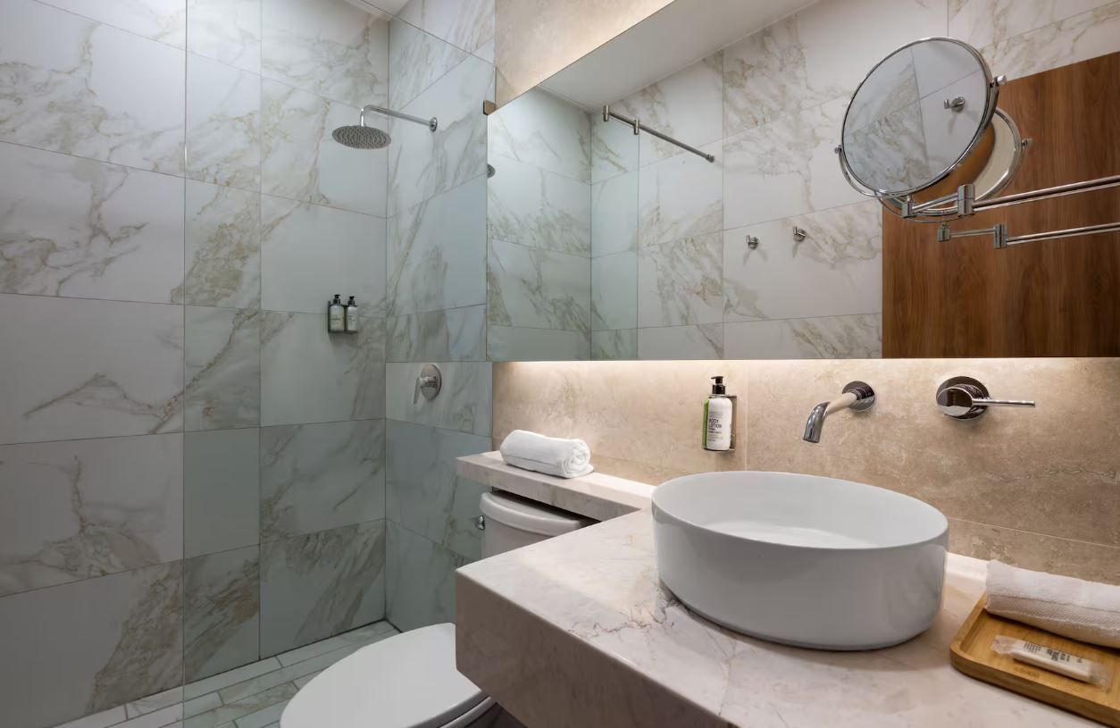Un baño moderno con ducha de azulejos de mármol, lavabo redondo y tocador de madera.