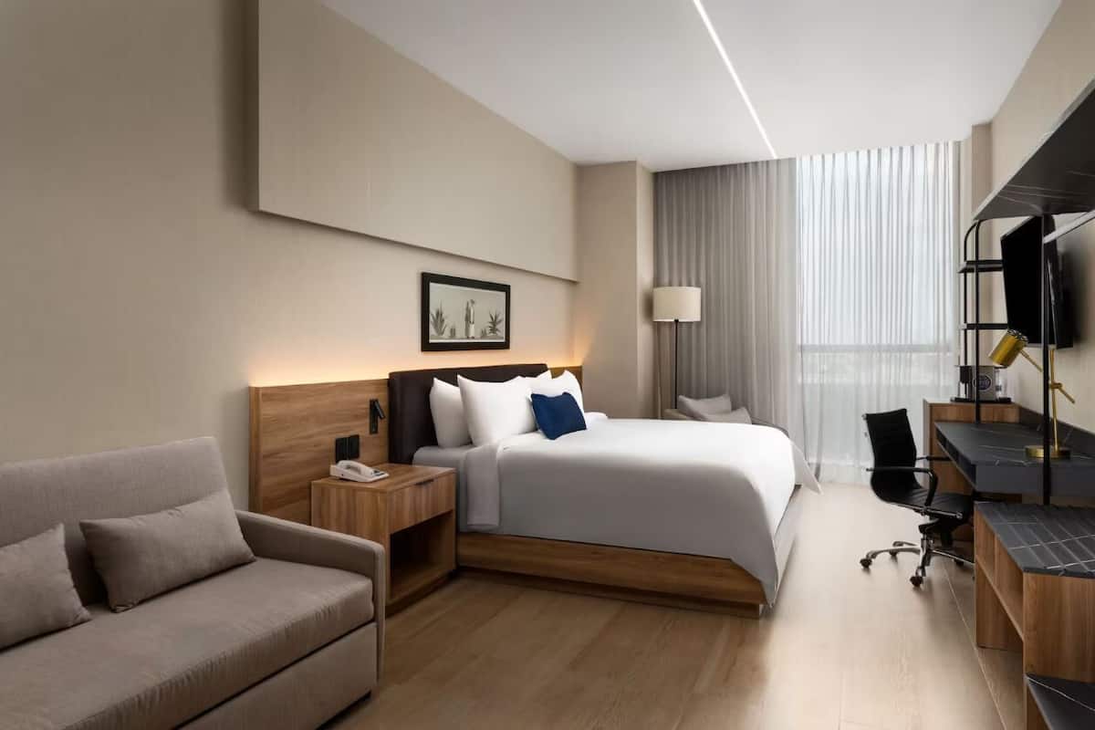 A modern hotel room with a bed, a desk, a chair, a sofa, and a TV.