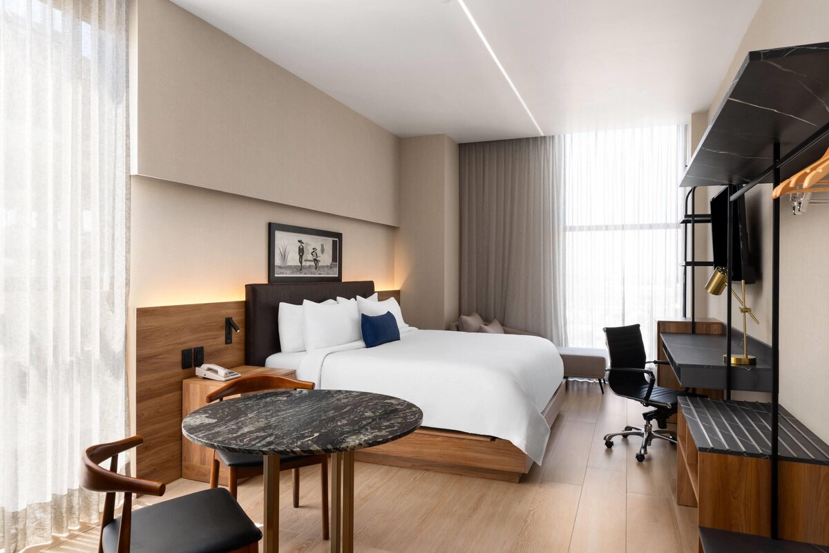 Une chambre d’hôtel moderne équipée d’un lit, d’un bureau, d’une chaise, d’une petite table et d’un téléviseur.