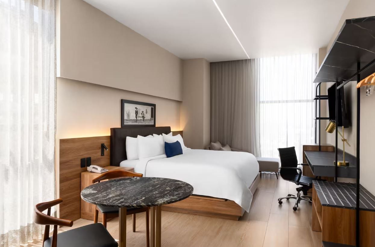 A modern hotel room with a large bed, a desk, a chair, a small table, and a television.