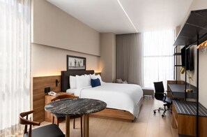 A modern hotel room with a large bed, a desk, a chair, a small table, and a television.