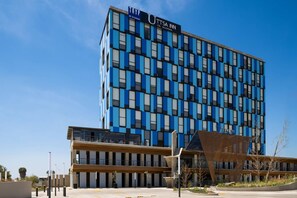 Un hotel moderno con fachada azul y blanca, un letrero llamativo que dice 'UTTSA INN', y un diseño arquitectónico distintivo con techo inclinado y una gran estructura de madera al frente.