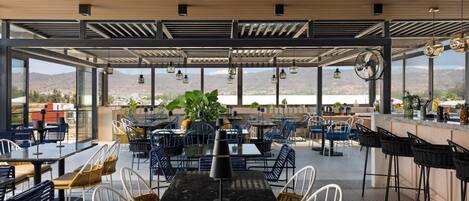 An outdoor dining area with tables and chairs, large windows, and a view of the landscape.