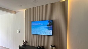 A modern living room with a flat-screen TV mounted on a textured wall, a black entertainment unit below, and a wooden floor. - Peaceful Apartment in Palmas del Mar (Palmas del Mar)