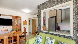 Sala de estar com sofá verde, mesa de jantar de madeira, cadeiras, televisão e área de cozinha com geladeira e armários.