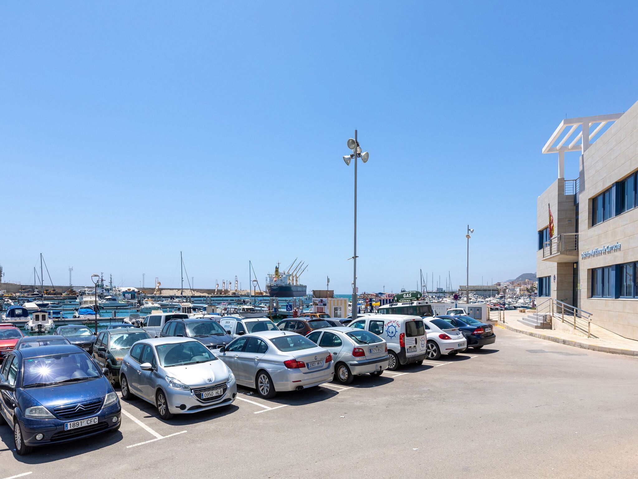 Un parking avec plusieurs voitures, un grand bâtiment avec une enseigne, et un port de plaisance en arrière-plan.