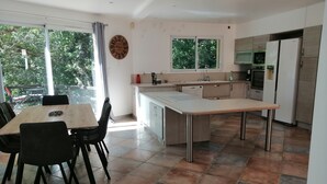 A modern kitchen with a dining area, large windows, and a view of greenery outside.