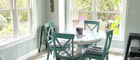 A dining area with a round table and four chairs, large windows with a view of the outdoors, and a ceiling vent.