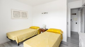 Una habitación con dos camas individuales, una mesita de noche pequeña con una lámpara y una puerta que da a otra habitación.