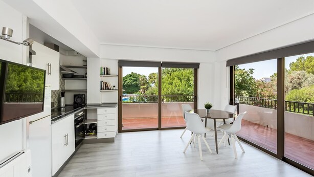 Cocina moderna con armarios blancos, encimera negra y horno integrado. Hay un comedor con mesa y sillas blancas, y un balcón con vistas a árboles y una piscina.