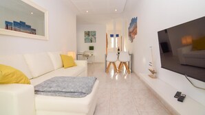 Una sala de estar moderna con un sofá blanco, una manta gris y un cojín amarillo. Hay un comedor con dos sillas blancas y una mesa. Un televisor de pantalla plana grande está montado en la pared.