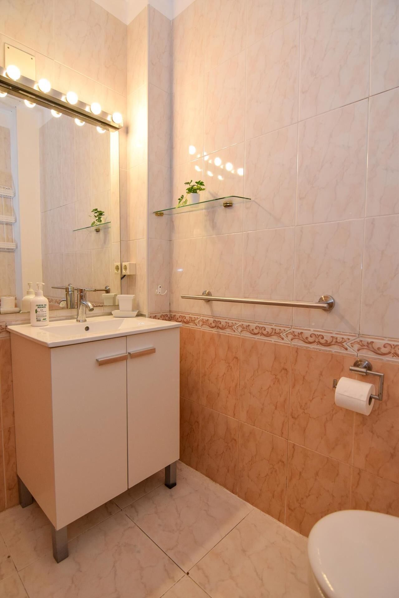 A modern bathroom with a vanity, sink, and mirror with lights. There is a towel rack, a toilet, and a glass shelf with a plant.