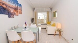 Un moderno salón con una mesa de comedor de cristal, sillas blancas, un sofá blanco con cojines amarillos, una mesita de cristal y un televisor montado en la pared.