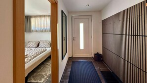 Ein schmaler Flur mit einem Bett links, einer Tür am Ende und einer Wand mit vertikalen Lamellen rechts.