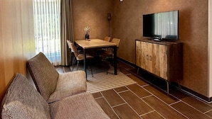 Ein Hotelzimmer mit einer Couch, einem Esstisch, Stühlen, einem Fernseher auf einer Holzkommode und einem Fenster mit Vorhängen.