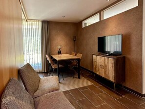 Habitación de hotel con sofá, mesa de comedor, sillas, televisor sobre un mueble de madera y una ventana con cortinas.
