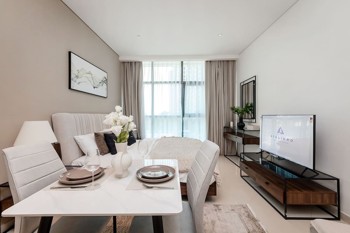 A modern hotel room with a bed, a dining table set with plates and glasses, a TV, and a large window with curtains.