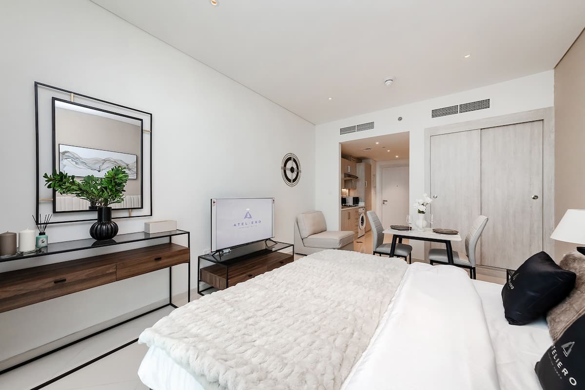 A modern hotel room with a bed, a desk with a computer, a chair, a small table, and a wardrobe.