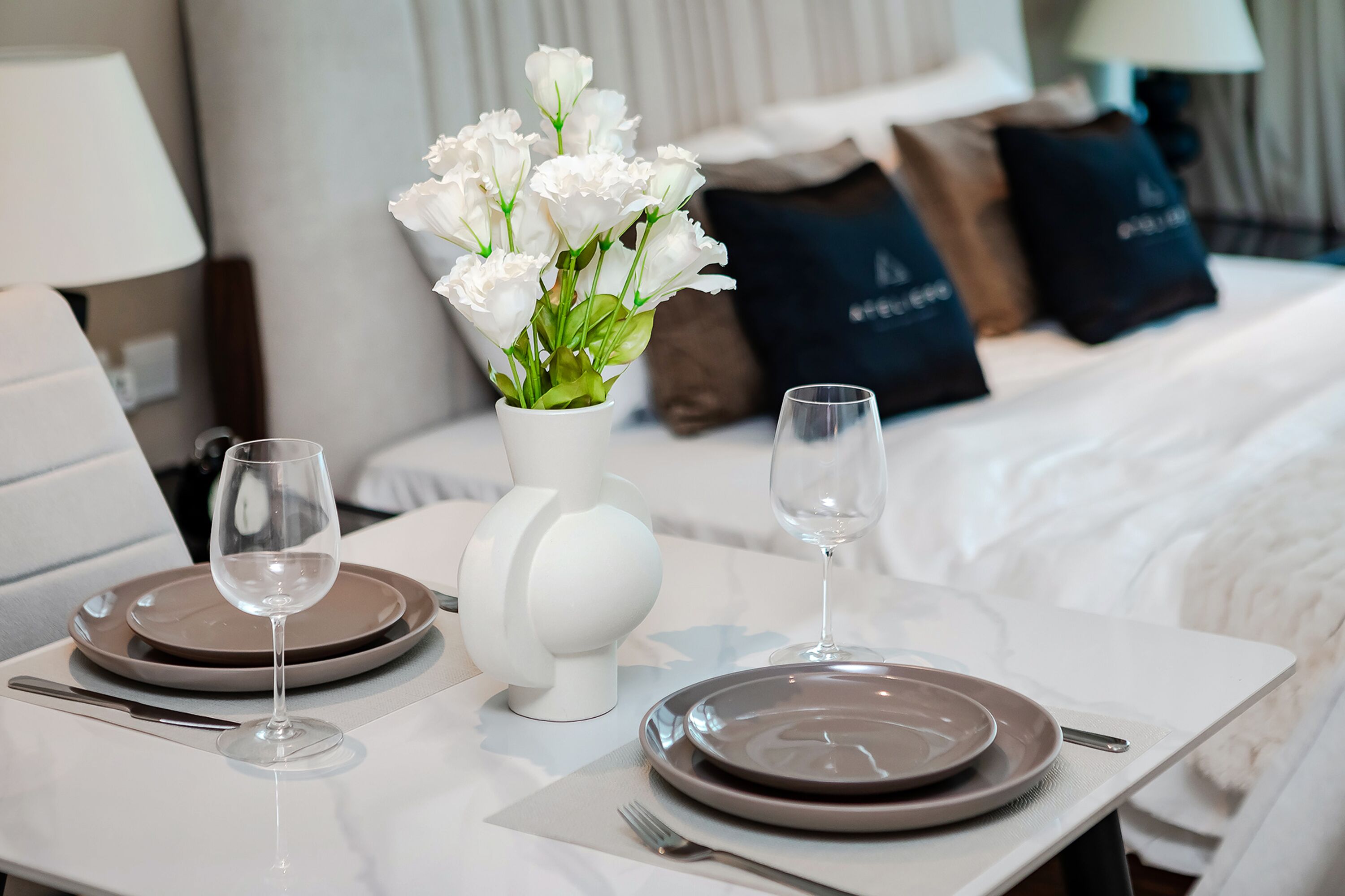 Uma mesa de jantar bem posta com dois pratos, talheres e copos de vinho, um vaso de flores brancas, e uma cama com almofadas ao fundo.