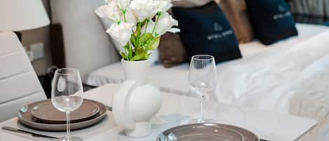 Uma mesa de jantar bem posta com dois pratos, talheres e copos de vinho, um vaso de flores brancas, e uma cama com almofadas ao fundo.