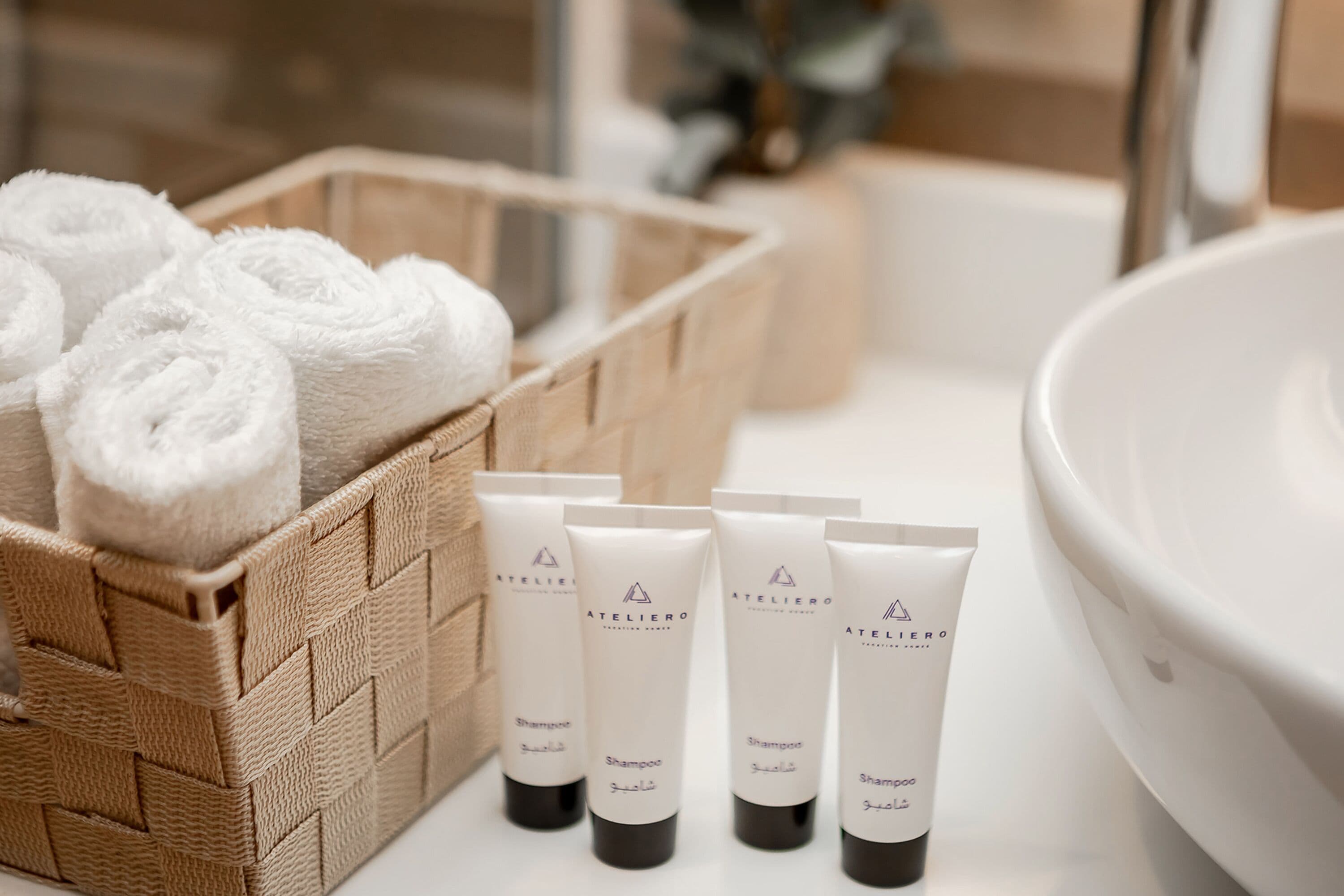 A bathroom with a large white sink, a wicker basket with rolled towels, and four white toiletry bottles with black caps.
