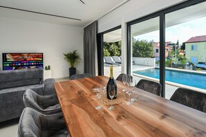 Una sala de estar moderna con una mesa de comedor de madera, un sofá gris y una gran pantalla de televisión. Se ve una piscina a través de los amplios ventanales.