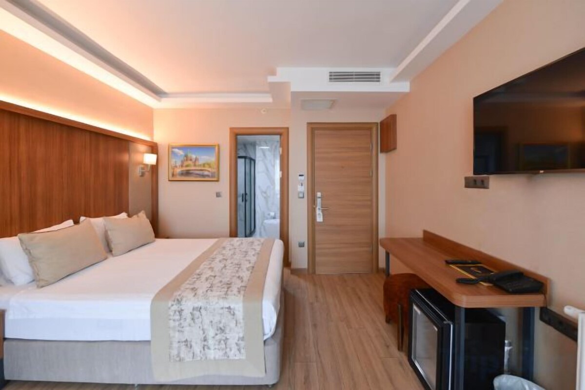 A hotel room with a large bed, a desk, a TV, and a door leading to another room.
