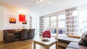 A modern living room with a wooden floor, a sofa, a red chair, a dining area with a table and chairs, and large windows with patterned curtains. - Apartment in Leogang near Ski Slopes (Leogang)