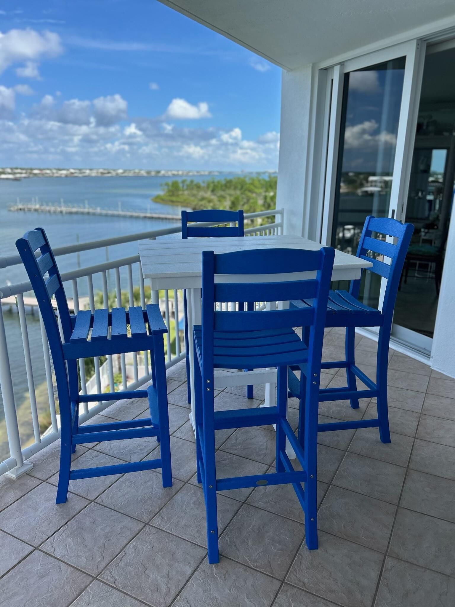 Ein Balkon mit Tisch und zwei blauen Stühlen, der auf ein Gewässer und eine entfernte Küste blickt.