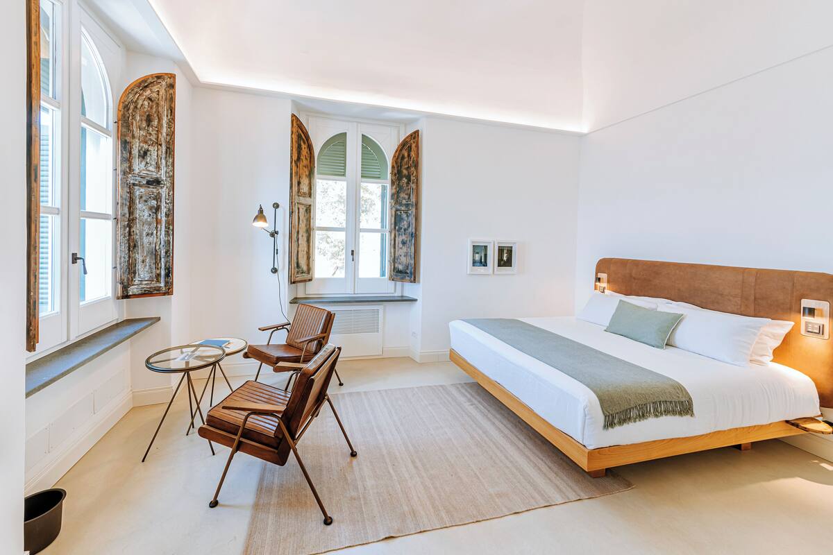 Quarto com uma cama grande, uma mesinha, duas cadeiras e uma janela com portadas.