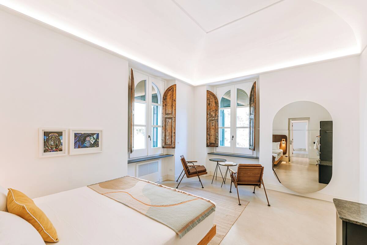 Um quarto moderno com uma cama grande, uma mesa redonda pequena e duas cadeiras. Há janelas amplas com arcos e um espelho na parede.
