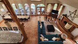 A spacious living area with a large sofa, a dining table, and a fireplace. There are multiple windows providing natural light and a view of the outdoors. - Perfect Summer Lakefront Escape- Pool/Outdoor Kitchen & BBQ/Hot Tub/Fire Pit (Canyon Lake)