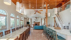 A spacious dining area with a long wooden table and chairs, a fireplace, and a staircase leading to an upper level. - Perfect Summer Lakefront Escape- Pool/Outdoor Kitchen & BBQ/Hot Tub/Fire Pit (Canyon Lake)