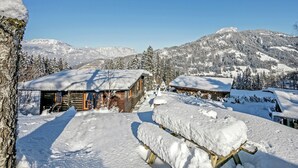 Ein mit Schnee bedecktes Blockhaus mit Holzfassade, umgeben von schneebedeckten Bäumen und Bergen im Hintergrund.