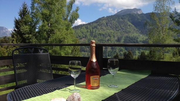 Ein Tisch mit einer Martini-Flasche, zwei Weingläsern und einer grünen Tischmatte, vor einer malerischen Bergkulisse.