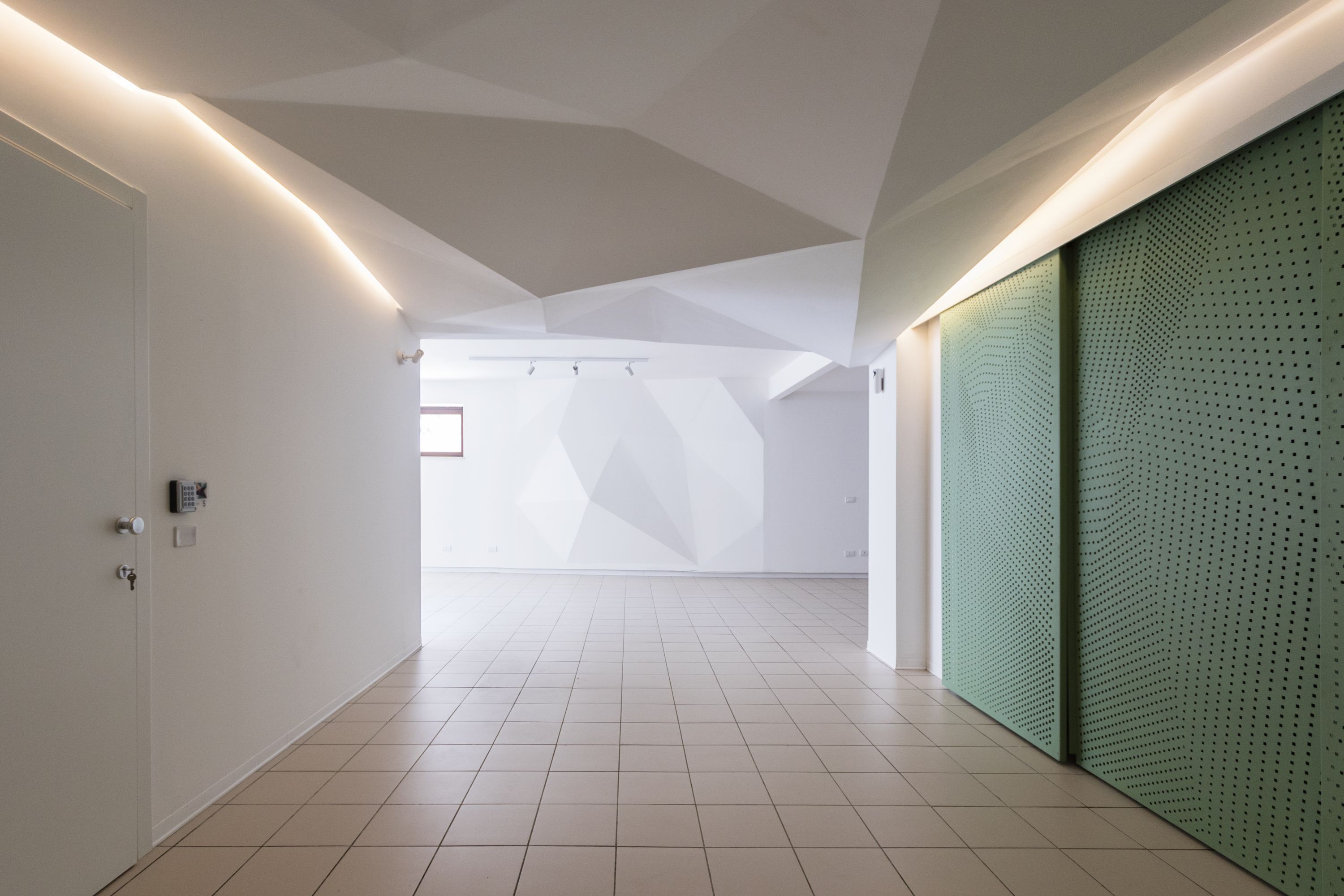 Un pasillo espacioso con un diseño geométrico en el techo, una pared verde perforada y una puerta con cerradura electrónica.