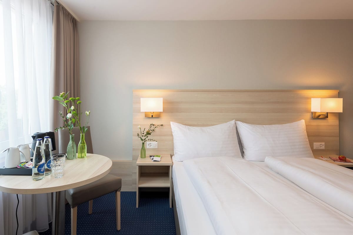 Habitación de hotel con cama, lámparas de noche, escritorio con silla, una mesita y un jarrón con flores.