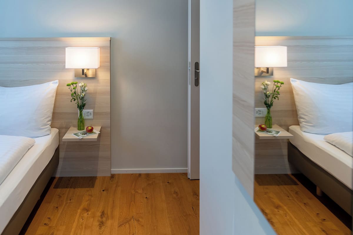 Habitación de hotel con suelo de madera, cama con ropa blanca, mesita de noche con lámpara y un jarrón con flores.