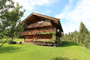 A two-story wooden chalet with a balcony, surrounded by a grassy area and trees. - Farmhouse in Hopfgarten near SkiWelt (Hopfgarten im Brixental)