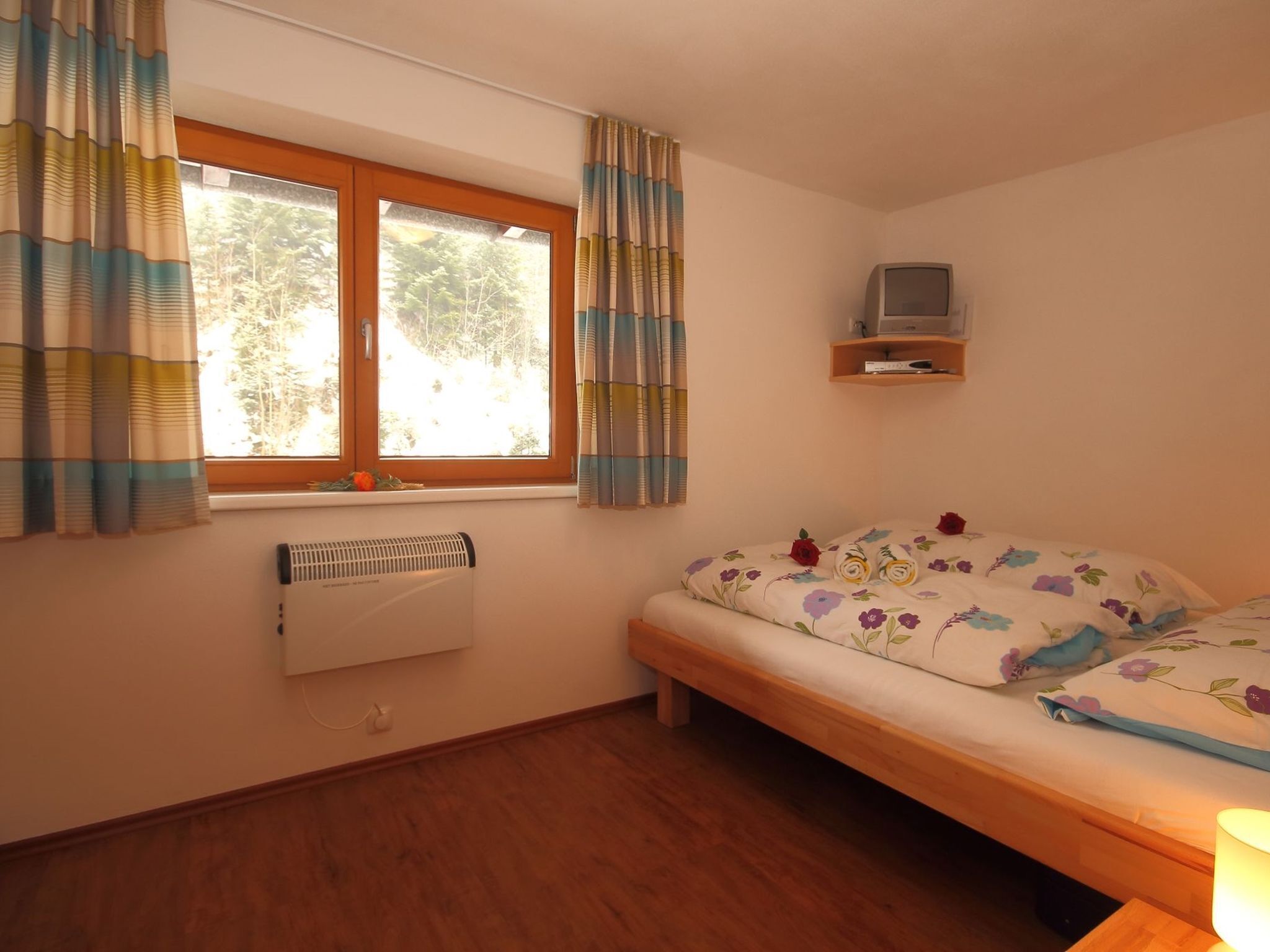 A bedroom with a bed, a window with curtains, a radiator, and a TV on a shelf.