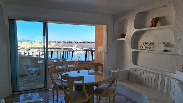 Une pièce avec une table ronde et des chaises, un canapé blanc et une porte vitrée donnant sur un balcon avec vue sur des bateaux et une marina.