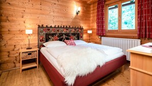 Un dormitorio de cabaña de madera con cama, dos mesitas de noche, una ventana con cortinas y un radiador.