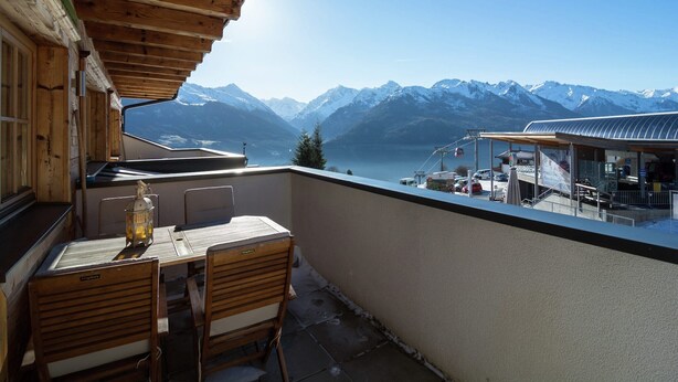 Ein Holzbalkon mit Tisch und Stühlen, mit Blick auf einen Bergsee und eine Seilbahn.