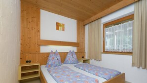 A room with a wooden bed, two pillows, a small shelf, a window with curtains, and a wall-mounted picture. - Apartment in Waidring near PillerseeTal Ski Area (Waidring)