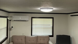 Un salon avec un canapé beige, un fauteuil foncé, un luminaire au plafond et une fenêtre avec des stores.