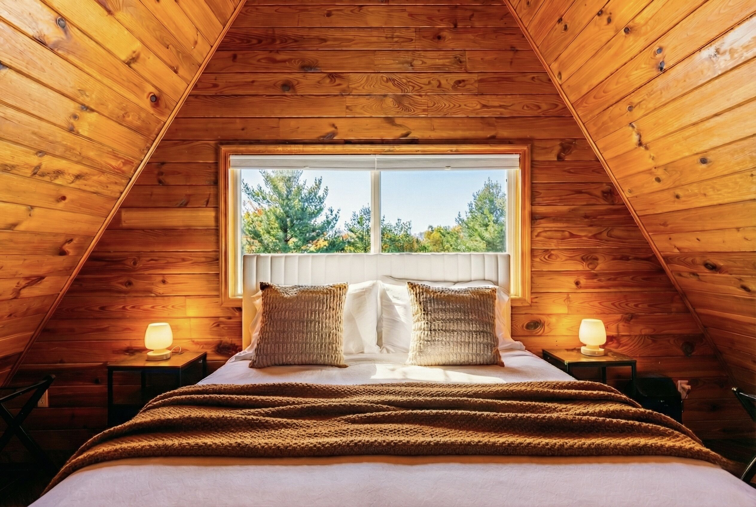 A bedroom with a sloped ceiling, a large window, a bed with pillows, and two bedside lamps.