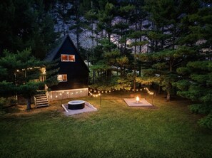 A cabin with a hot tub and a fire pit in the yard, surrounded by trees.